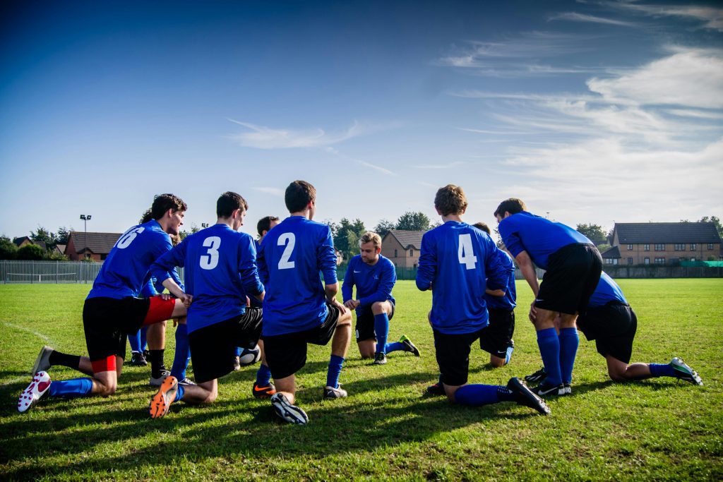 A group of players take a knee together on a football field, discussing team sports benefits